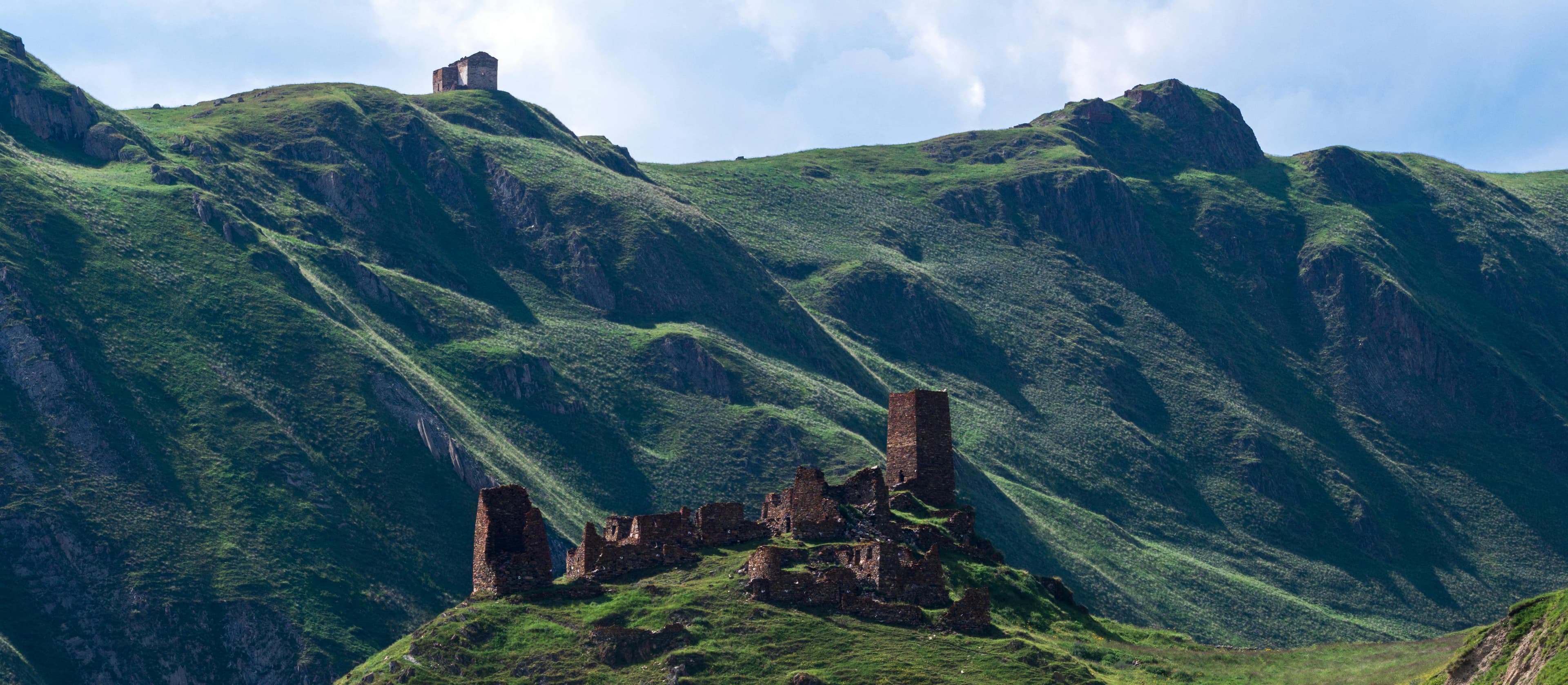 Ancient stone fortress ruins overlooking Truso Valley with dramatic Kazbegi mountain peaks and alpine meadows in background