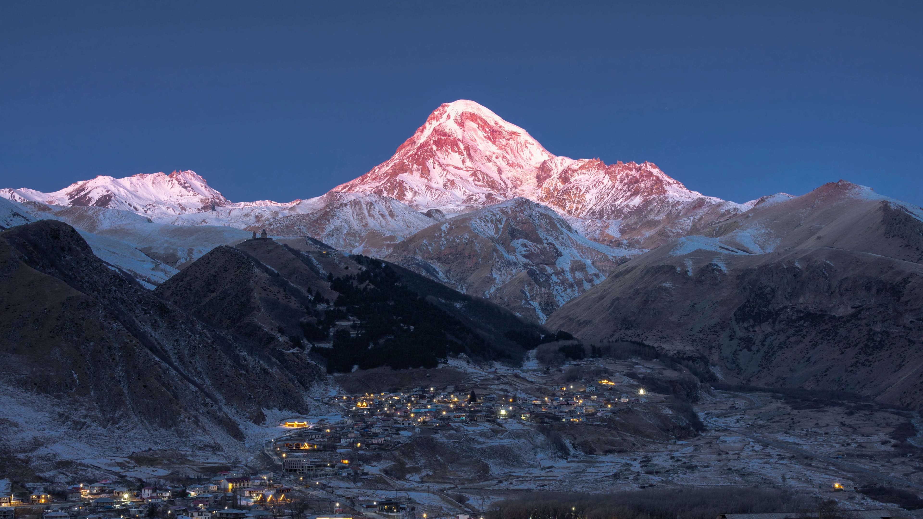 Dramatic sunset over Mount Kazbek with Gergeti Trinity Church silhouetted against golden sky in Georgian Caucasus Mountains